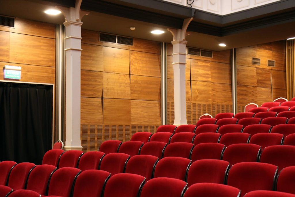 red-theater-chair-lot-near-white-concrete-pillars-269140 Interior of an empty auditorium with red seats and wooden panels, lit overhead.